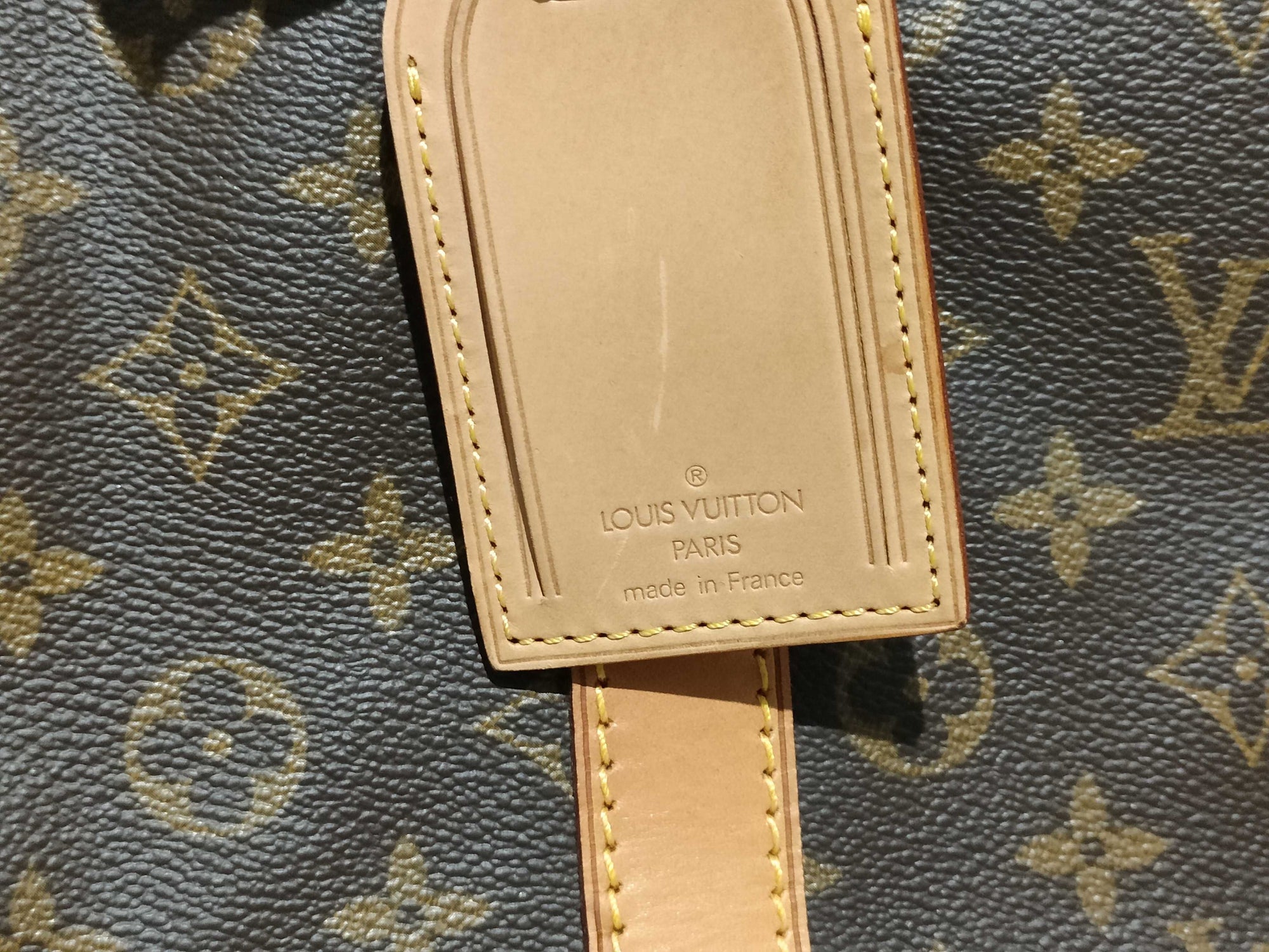 Close-up of a Louis Vuitton Keepall 55 luggage tag with 'Made in France' stamp on brown monogram coated canvas.